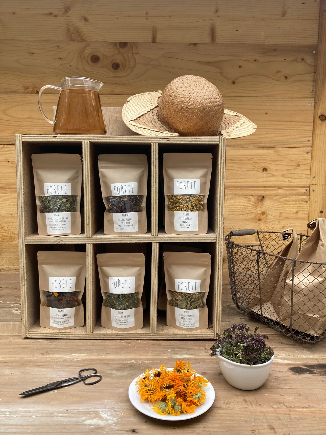 Les tisanes en vrac de la Forête dans leurs sachets kraft sur une étagère en bois, mon chapeau de jardin, un panier, des ciseau, une théière