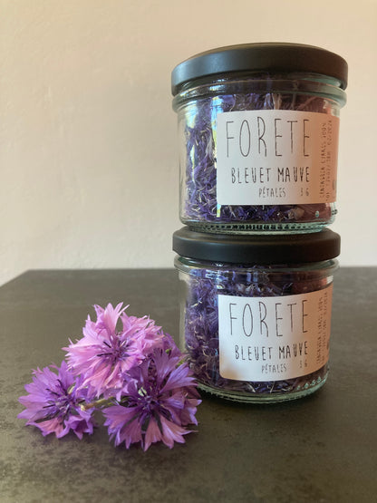 Deux pots en verre remplis de pétales de Bleuets mauves Forête accompagnés de 3 fleurs fraîches de Bleuets mauves posés les uns sur les autres.